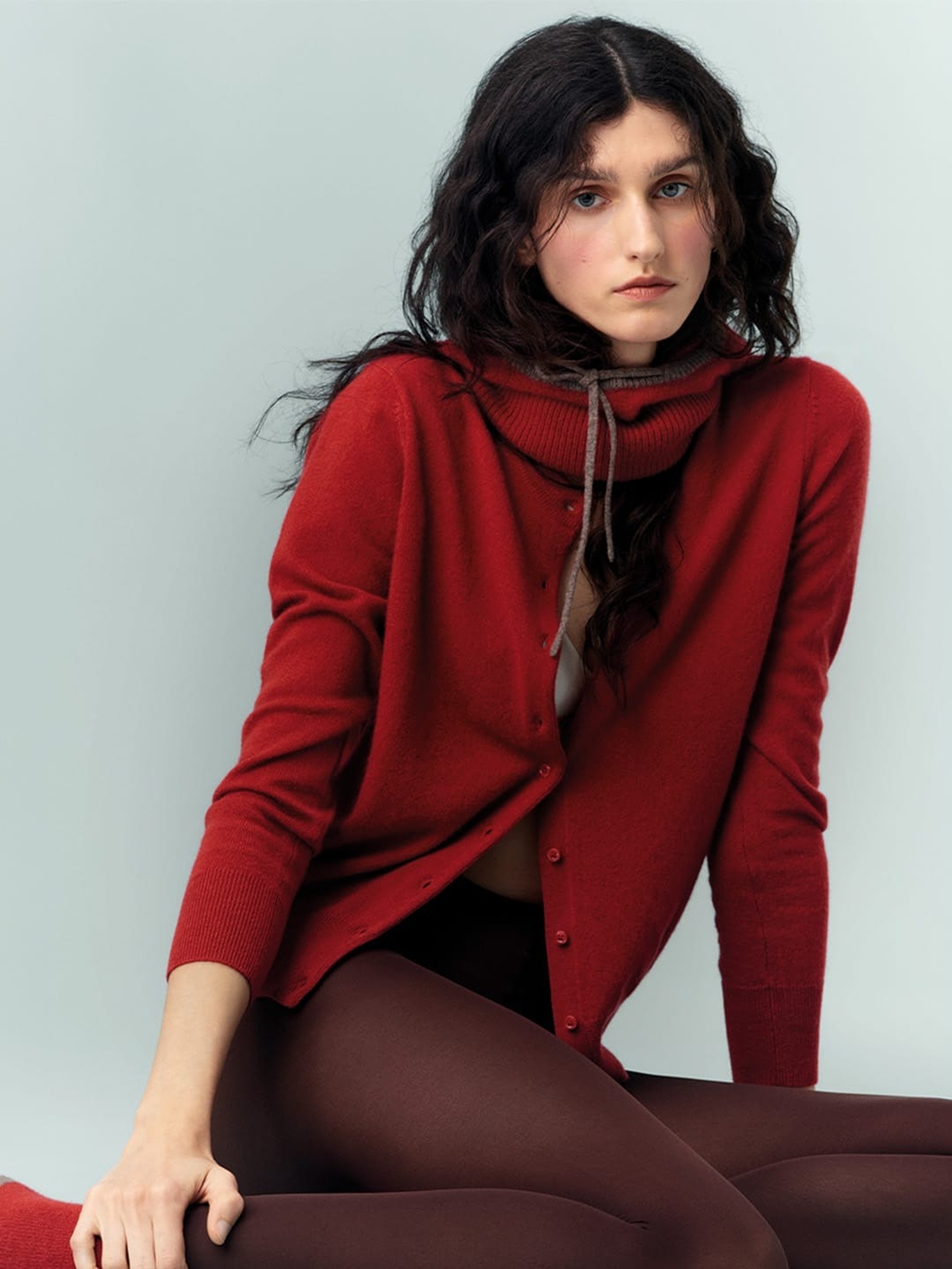 Model wearing a red cardigan and matching knit scarf with dark brown tights, seated against a soft light grey background